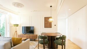 Moderni olohuone, jossa on pyöreä ruokapöytä, vihreäverhoillut tuolit, harmaa sohva, litteänäyttöinen televisio ja koristeellinen lamppu.