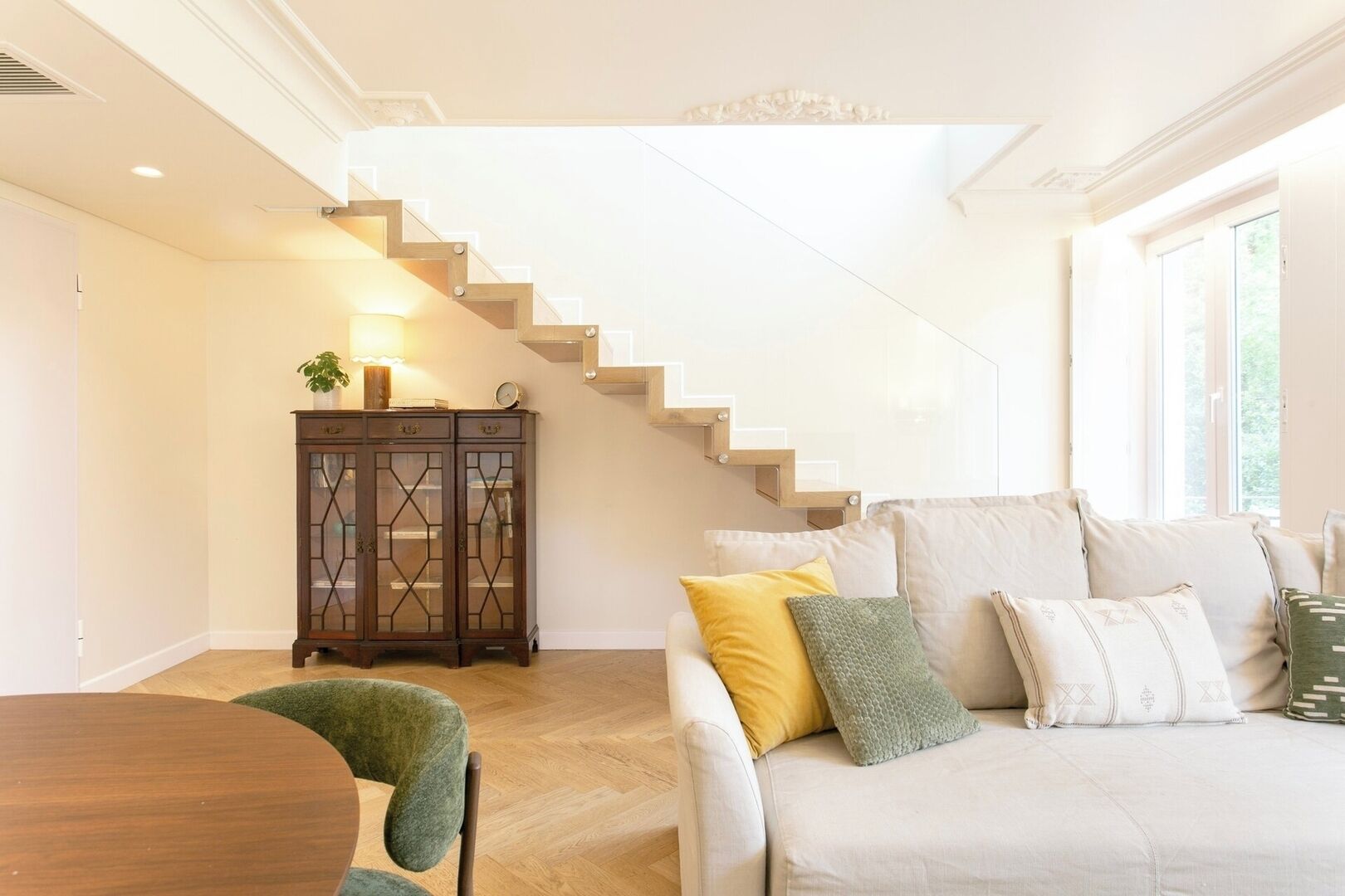 A modern living room with a staircase, a wooden cabinet, and a sofa with cushions.