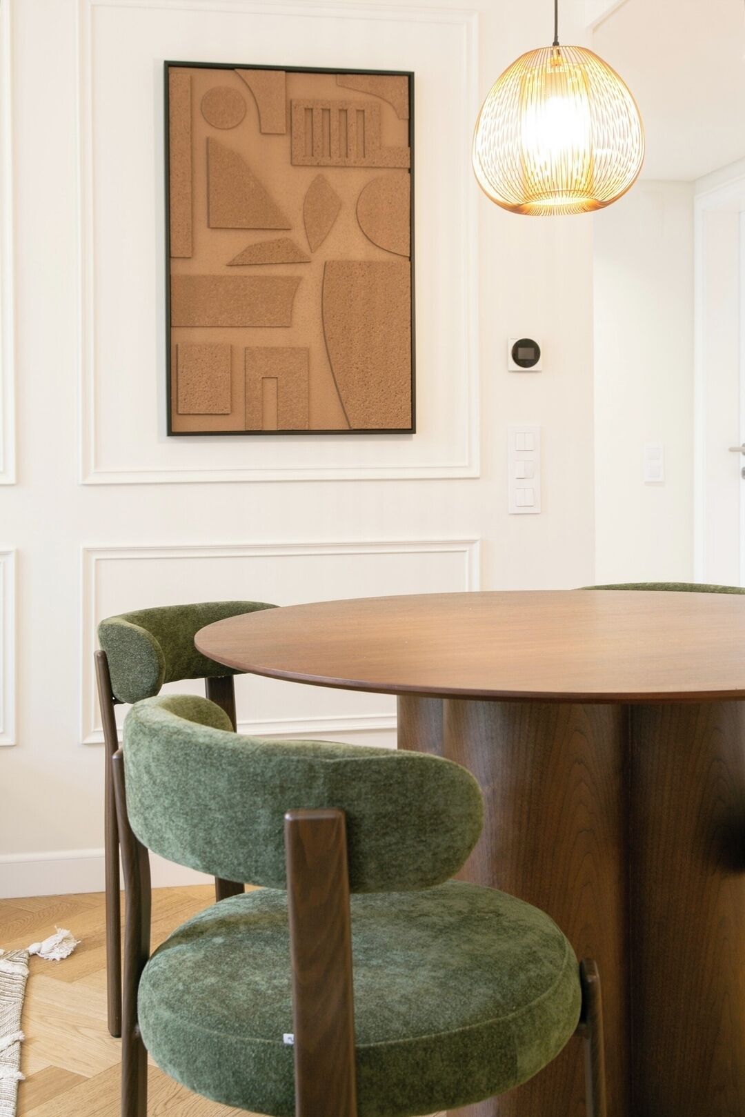 A dining area with a round wooden table and two green upholstered chairs. A textured pendant light hangs above the table.