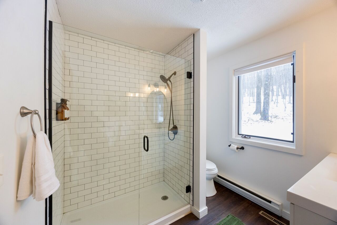 Une salle de bain moderne avec une douche à parois de verre, des murs recouverts de carreaux blancs, des toilettes et une fenêtre donnant sur un paysage enneigé.