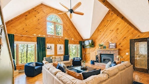 Un intérieur de chalet chaleureux avec un plafond de style industriel, une cheminée et une grande fenêtre donnant sur un paysage verdoyant.