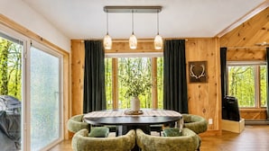 Un coin repas comprenant une table ronde, quatre chaises rembourrées, un vase avec une plante et des murs en bois dotés de grandes fenêtres.
