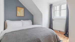 Une chambre avec un plafond en pente, un lit recouvert d’une couette blanche à motifs, une œuvre d’art encadrée accrochée au mur et une fenêtre avec des rideaux.