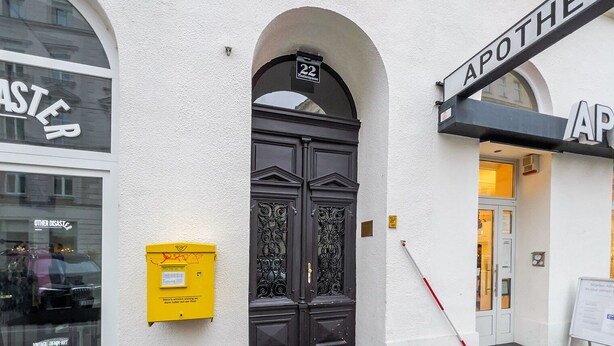 Un bâtiment doté d’une porte noire ornée, d’une boîte aux lettres jaune et d’une enseigne indiquant « APOTHEKE ».