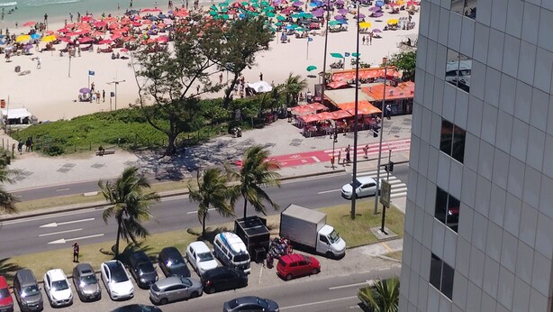 Une plage avec des parasols et des gens, une route avec des voitures, et un bâtiment au premier plan.