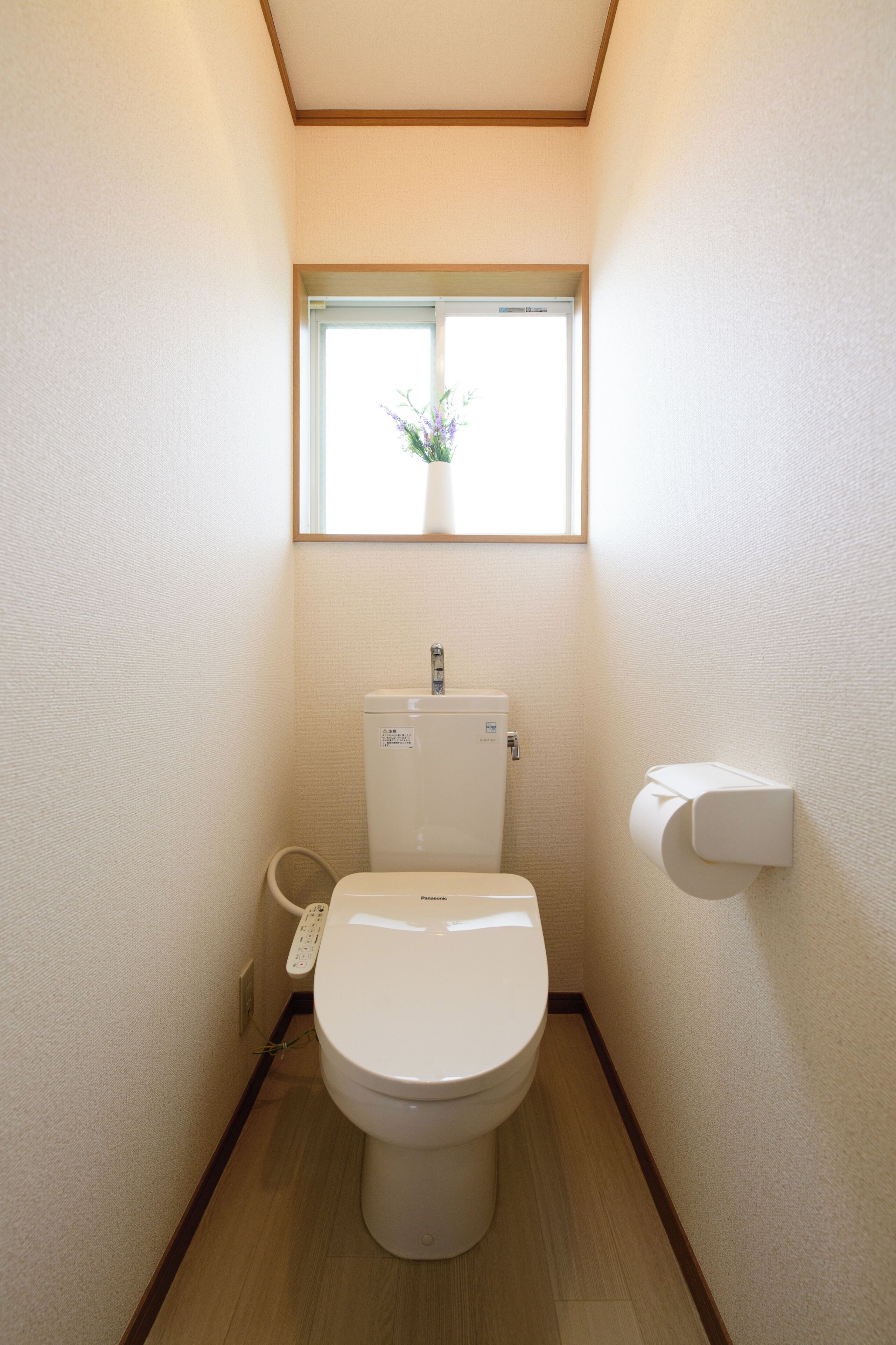 A compact bathroom with a toilet, a small window with a plant, and a wall-mounted toilet paper holder.