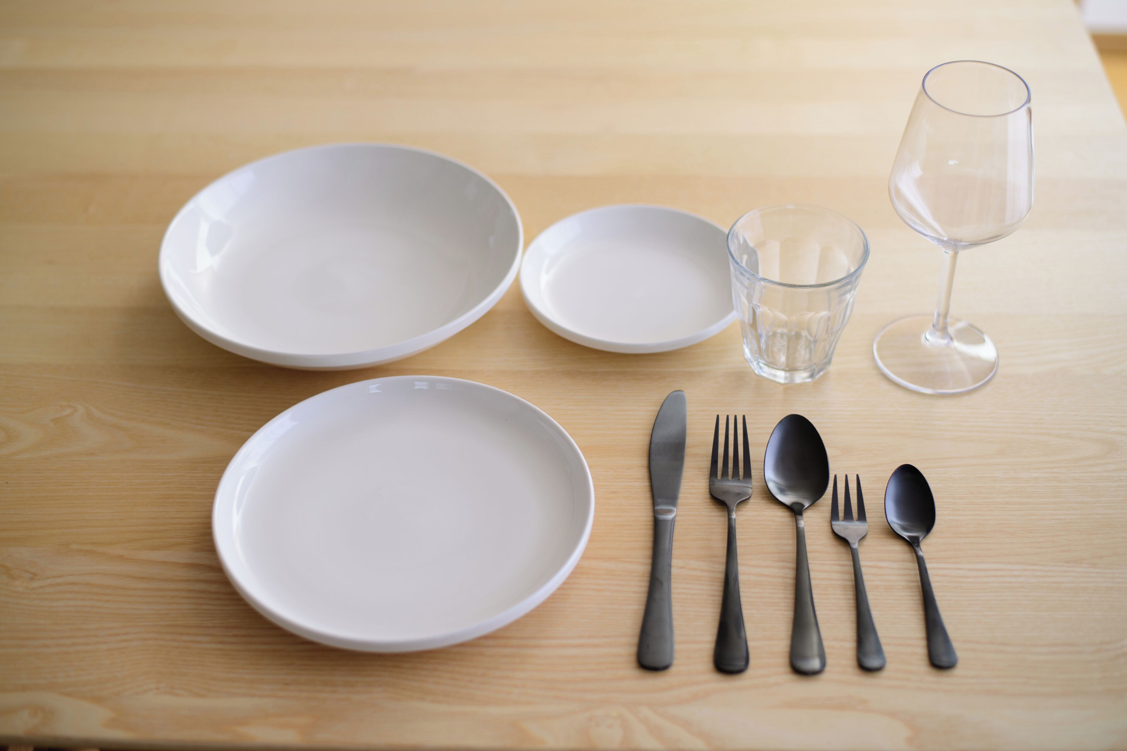 Una mesa de madera con cuatro platos blancos, un vaso, una copa de vino y cubiertos que incluyen un cuchillo, un tenedor, una cuchara y otra cuchara.