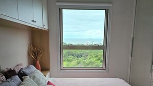 A bedroom with a large window, a bed with multiple pillows, and a view of greenery and buildings. - Sunrise apartment (Itajaí)