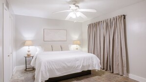 Un dormitorio con cama, dos lámparas de noche, ventilador de techo, una obra de arte enmarcada en la pared y una ventana con cortinas.