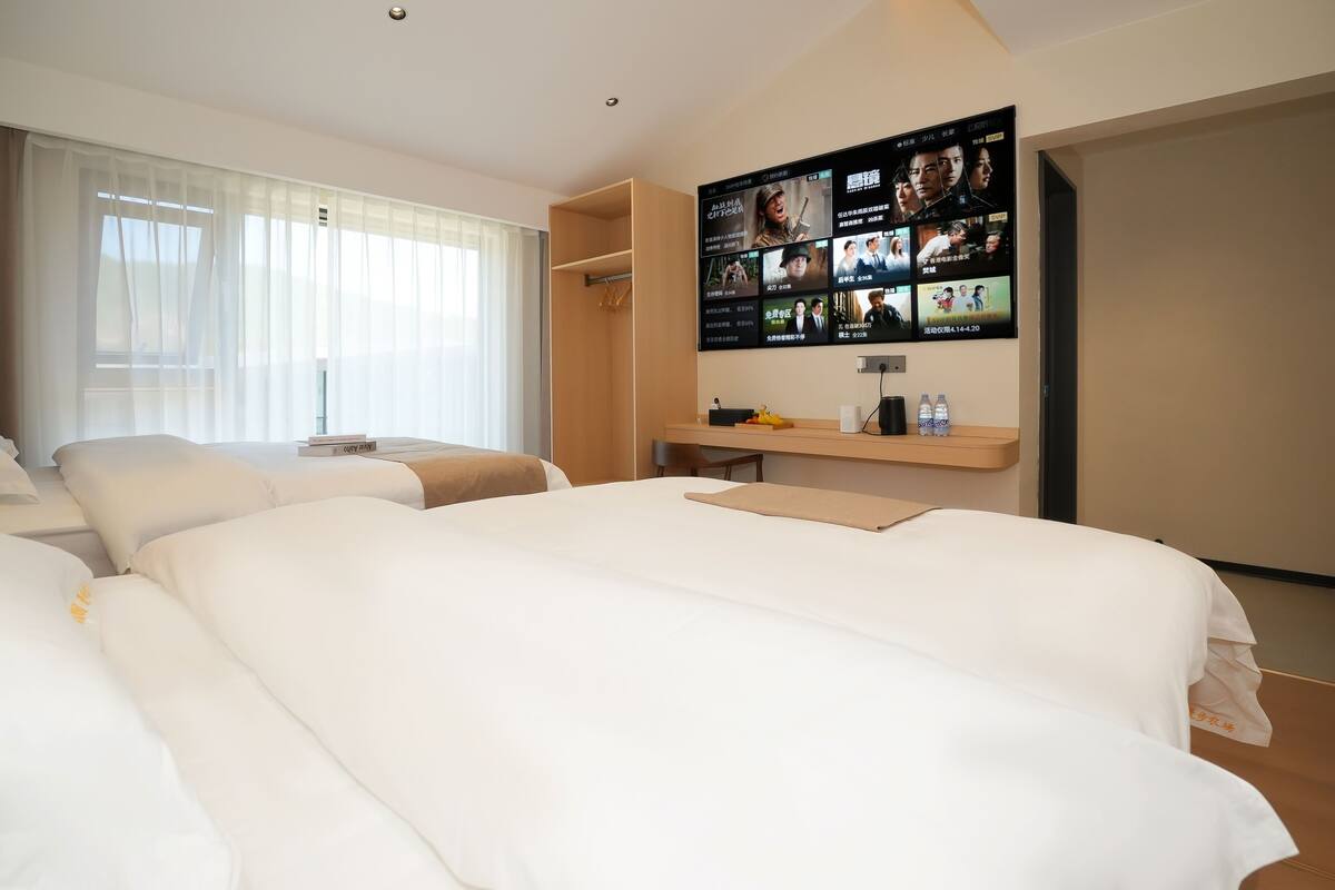 A hotel room with two beds, a TV wall, and a window with sheer curtains.