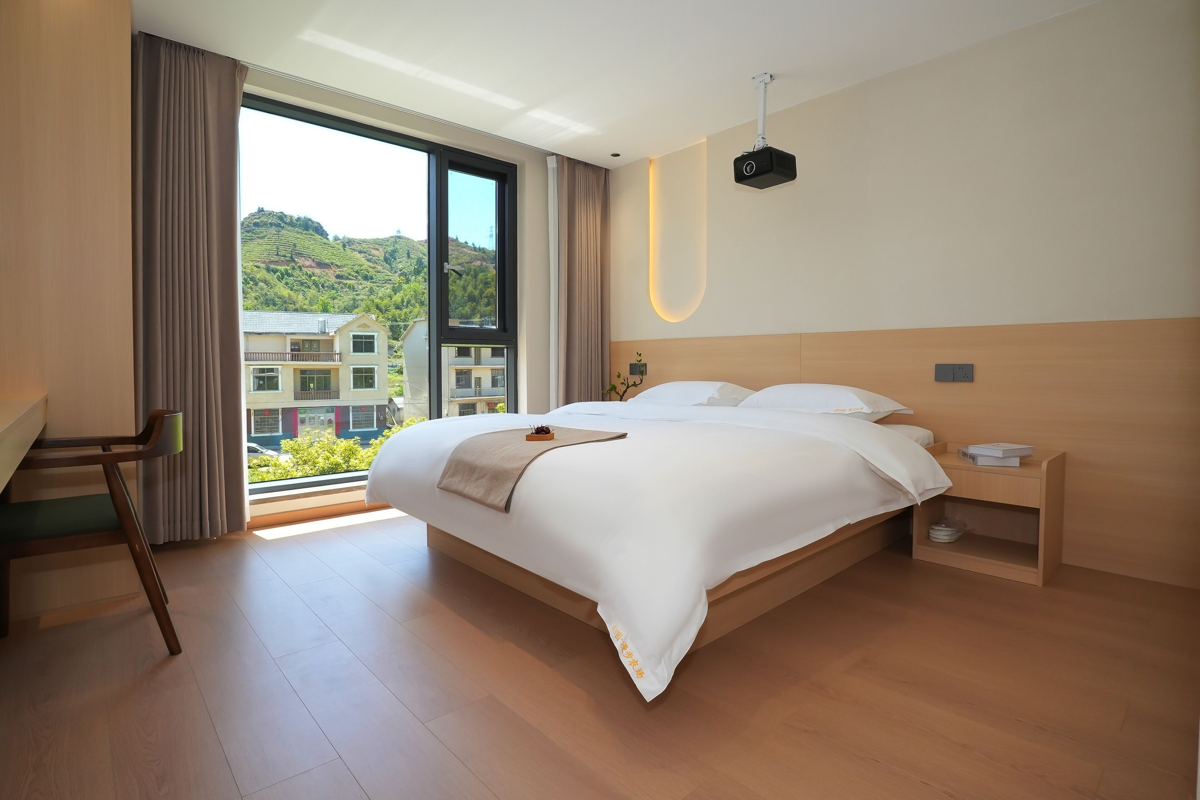 A modern hotel room with a large bed, a desk, and a view of a building and hills.