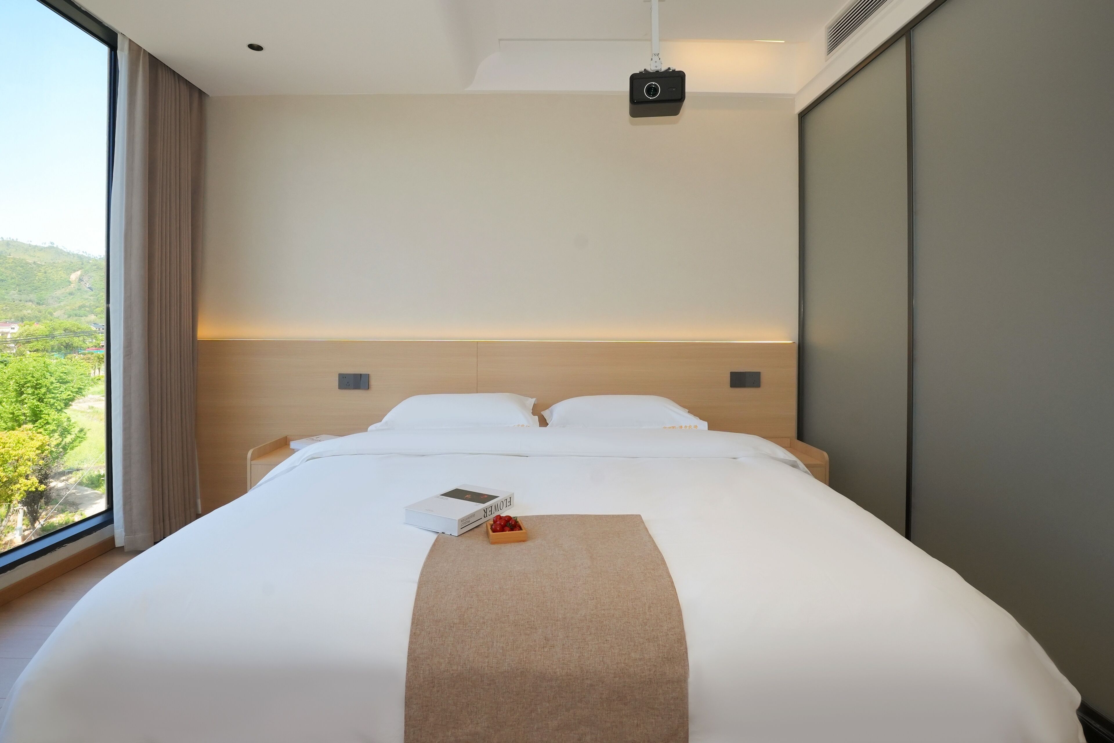 Habitación de hotel con una cama grande, vistas a zonas verdes, una mesita de noche con un libro y fruta, y una cámara en el techo.