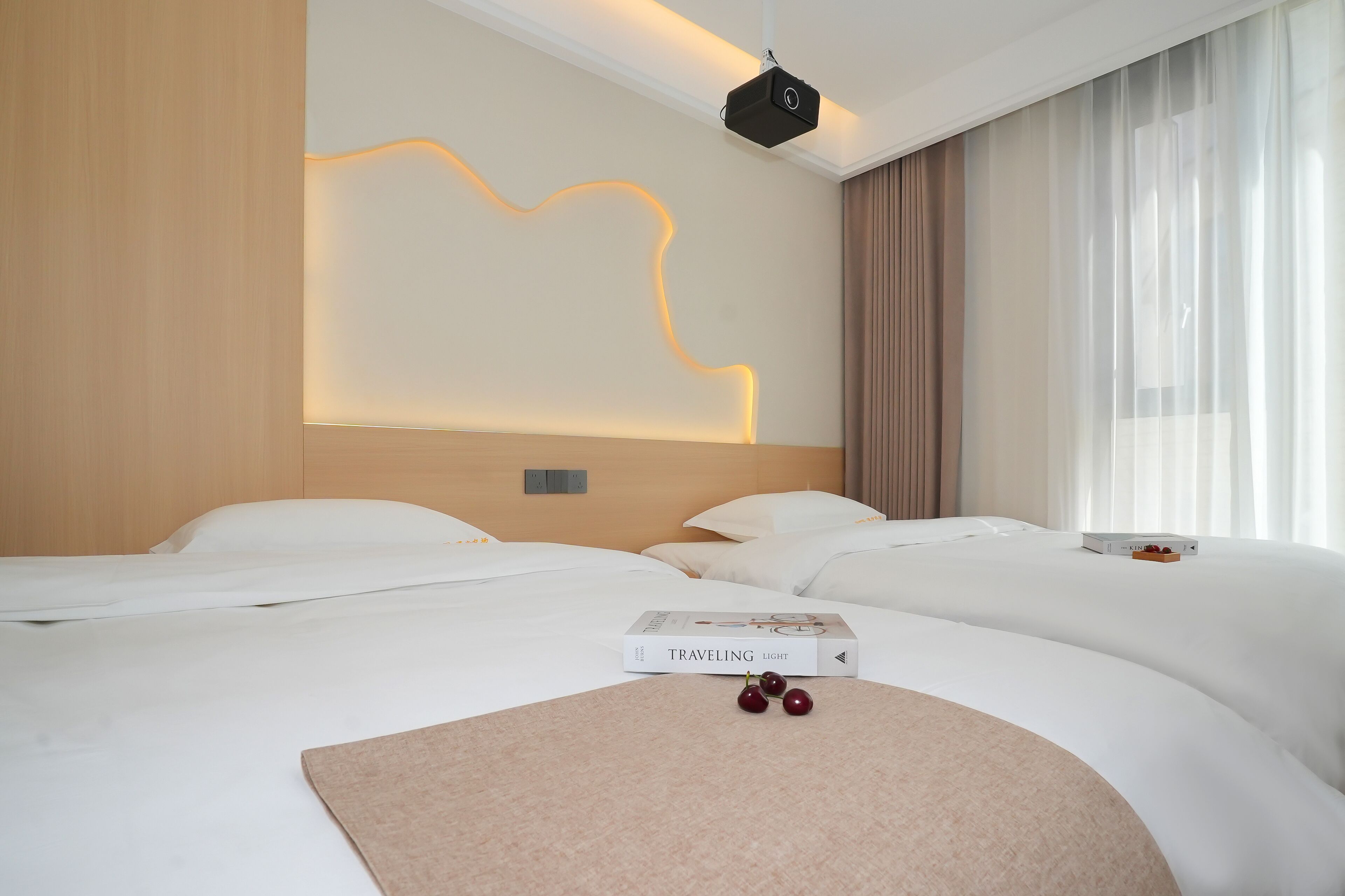 A hotel room with two beds, a wall-mounted projector, and a book titled "TRAVELING LIGHT" on the bed.