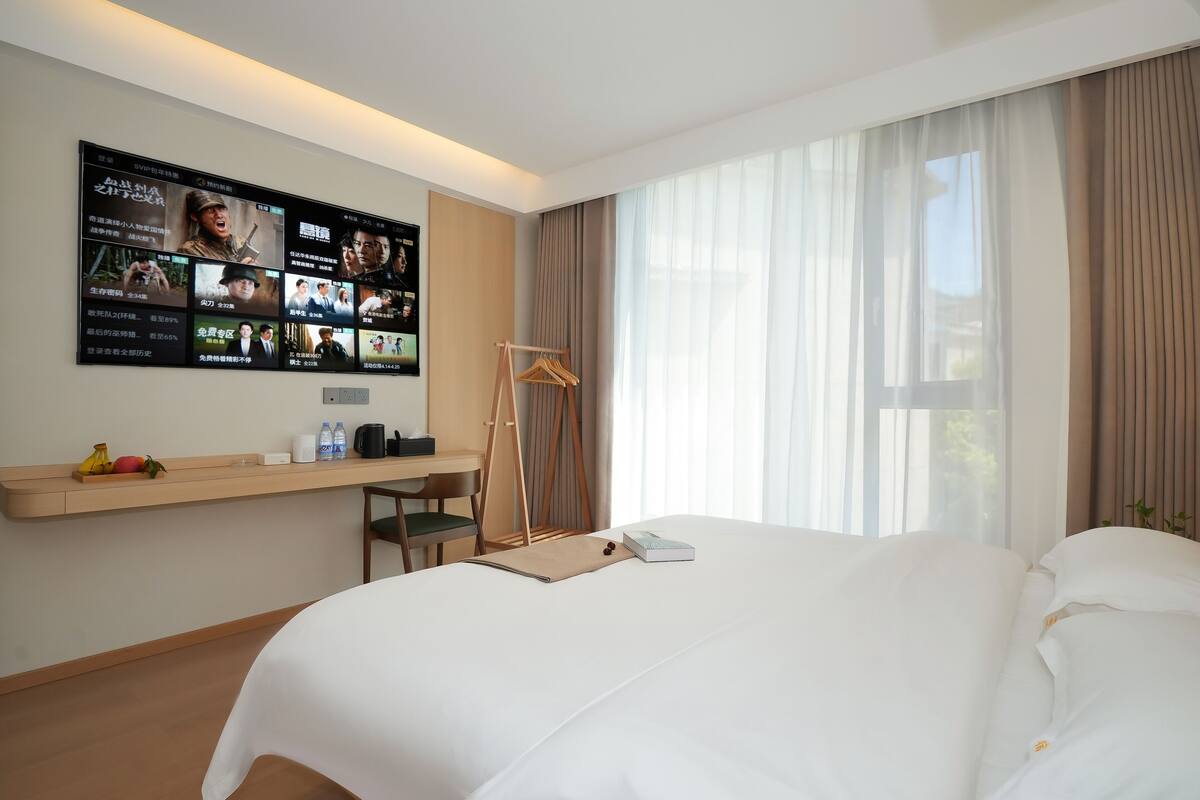 A hotel room with a large bed, a desk with a chair, a TV mounted on the wall displaying movie options, and a window with curtains.