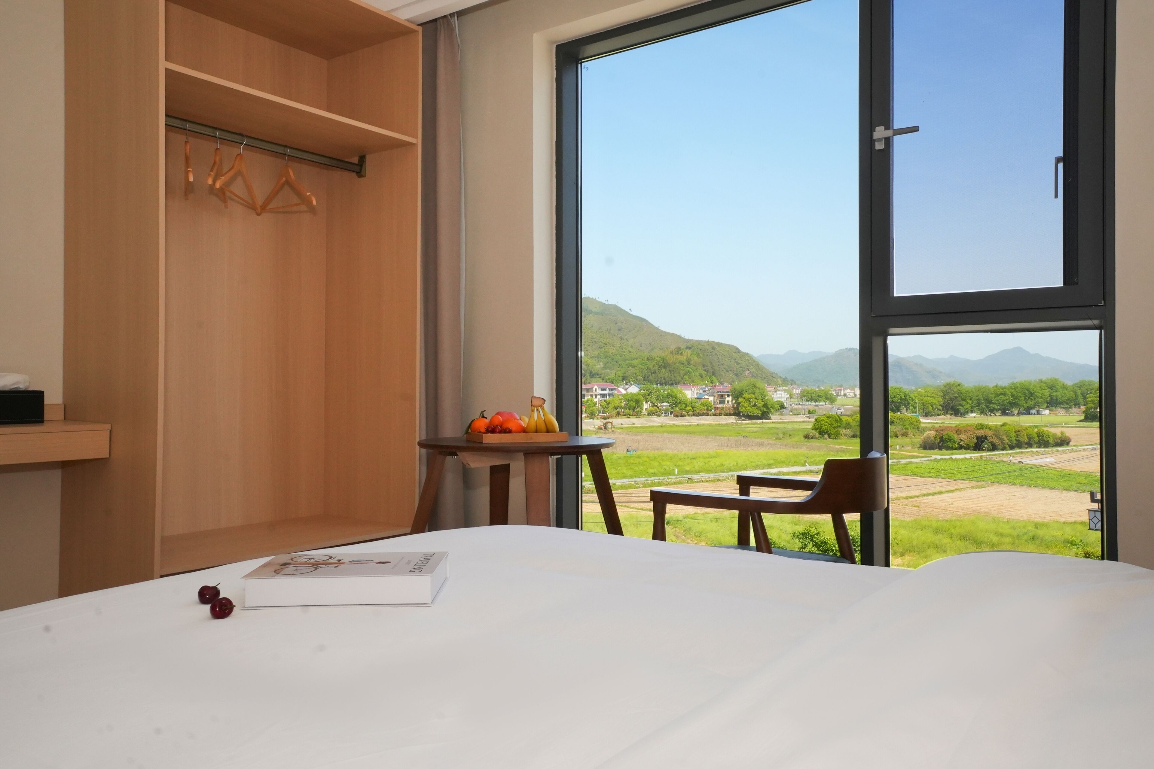 A hotel room with a large window, a bed, a small table with fruit, and a wooden wardrobe.