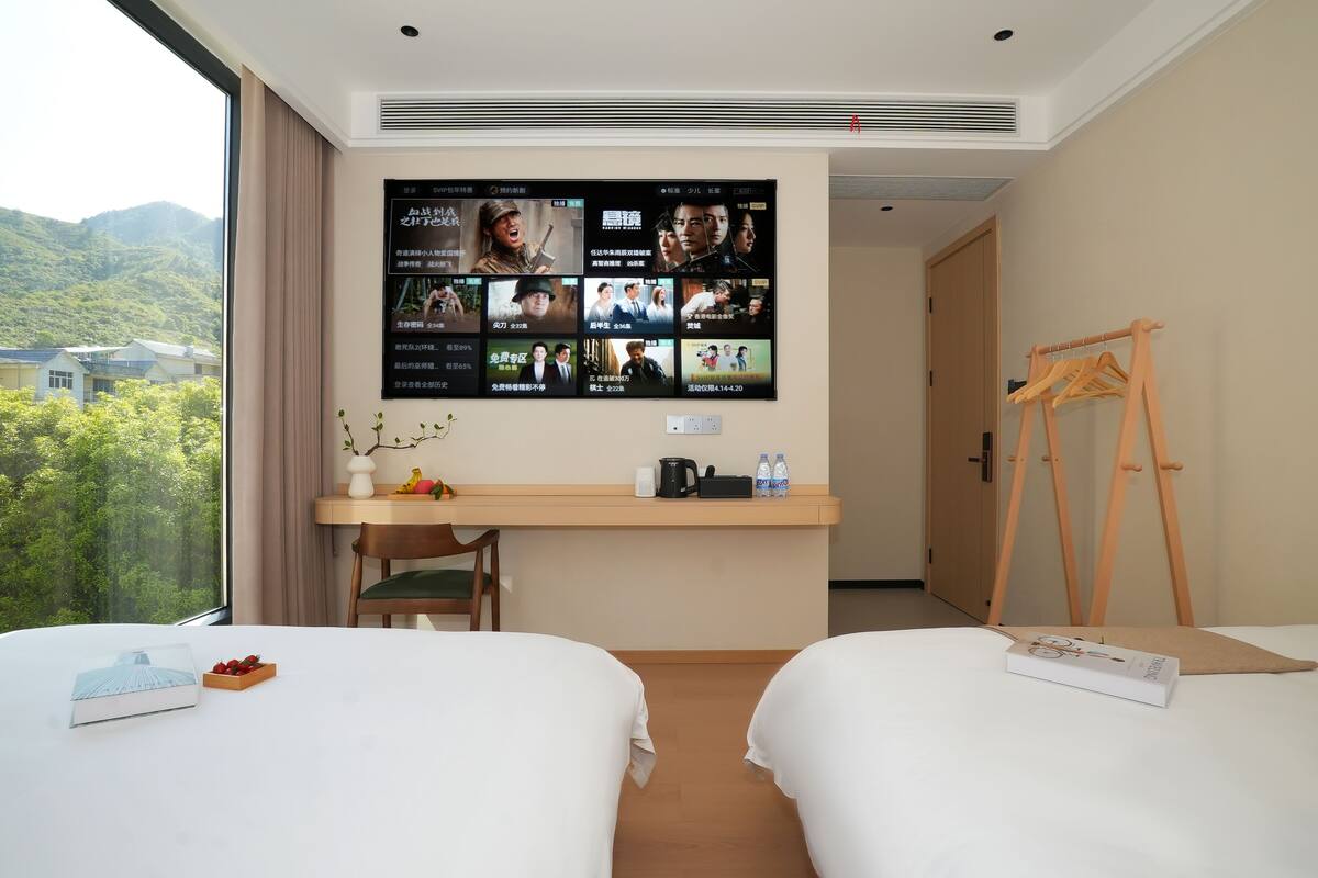 A hotel room with two beds, a desk with a flat-screen TV displaying movie posters, a view of greenery outside, and a wooden easel.