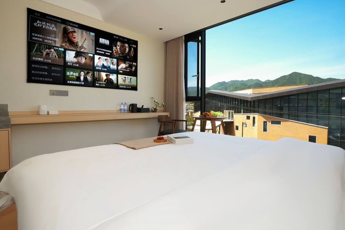 A hotel room with a large bed, a TV mounted on the wall displaying movie posters, a window with a view of mountains, and a small table with a plant and a book.