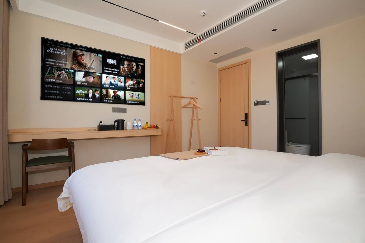 A hotel room with a large bed, a desk with a computer, a TV displaying movie posters, a wooden chair, and a wardrobe.