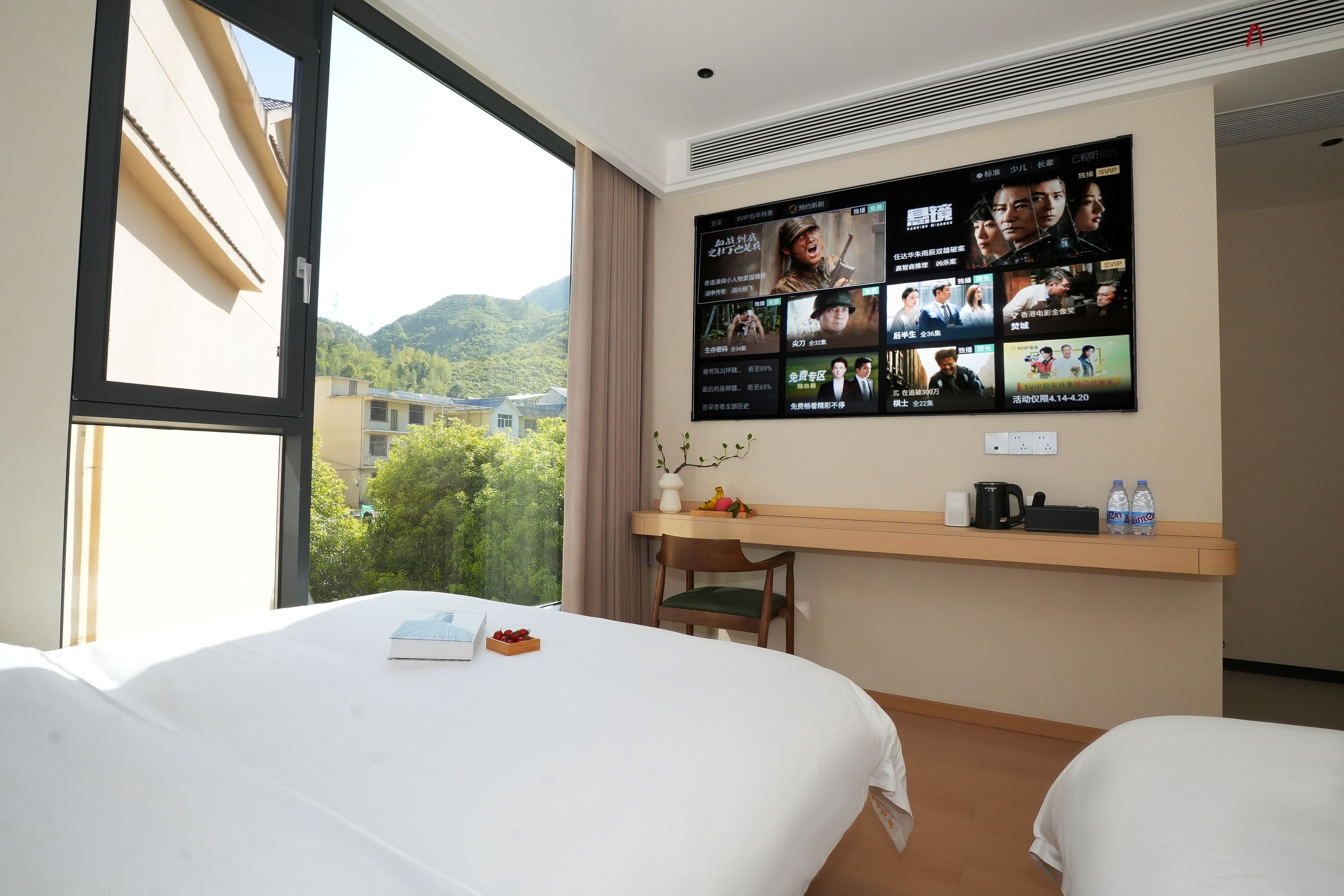 Hotelkamer met twee bedden, een aan de muur bevestigde televisie met filmopties, een bureau met stoel en uitzicht op groen buiten.