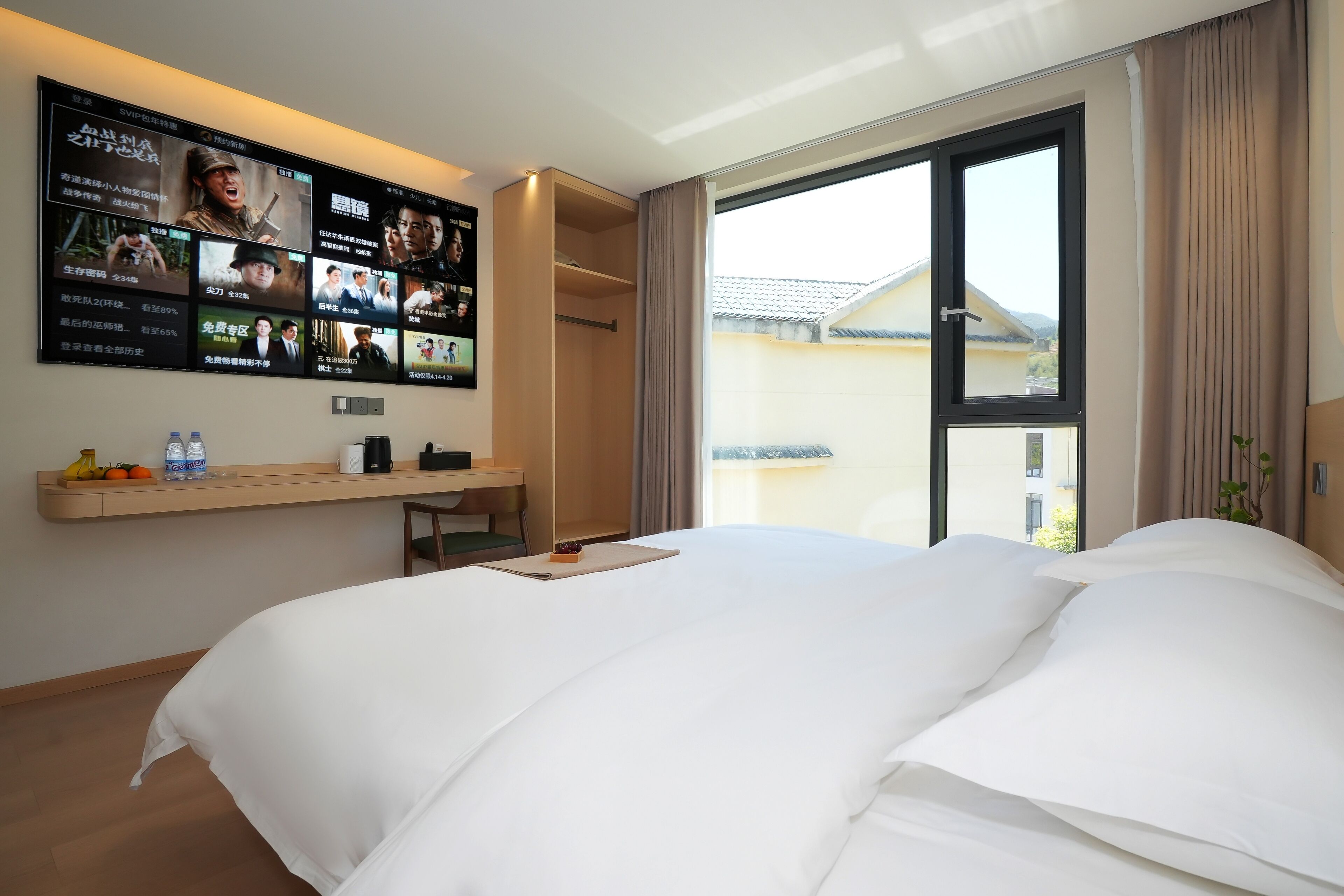 A hotel room with a large bed, a TV mounted on the wall displaying movie posters, a small desk with a chair, and a window with curtains.