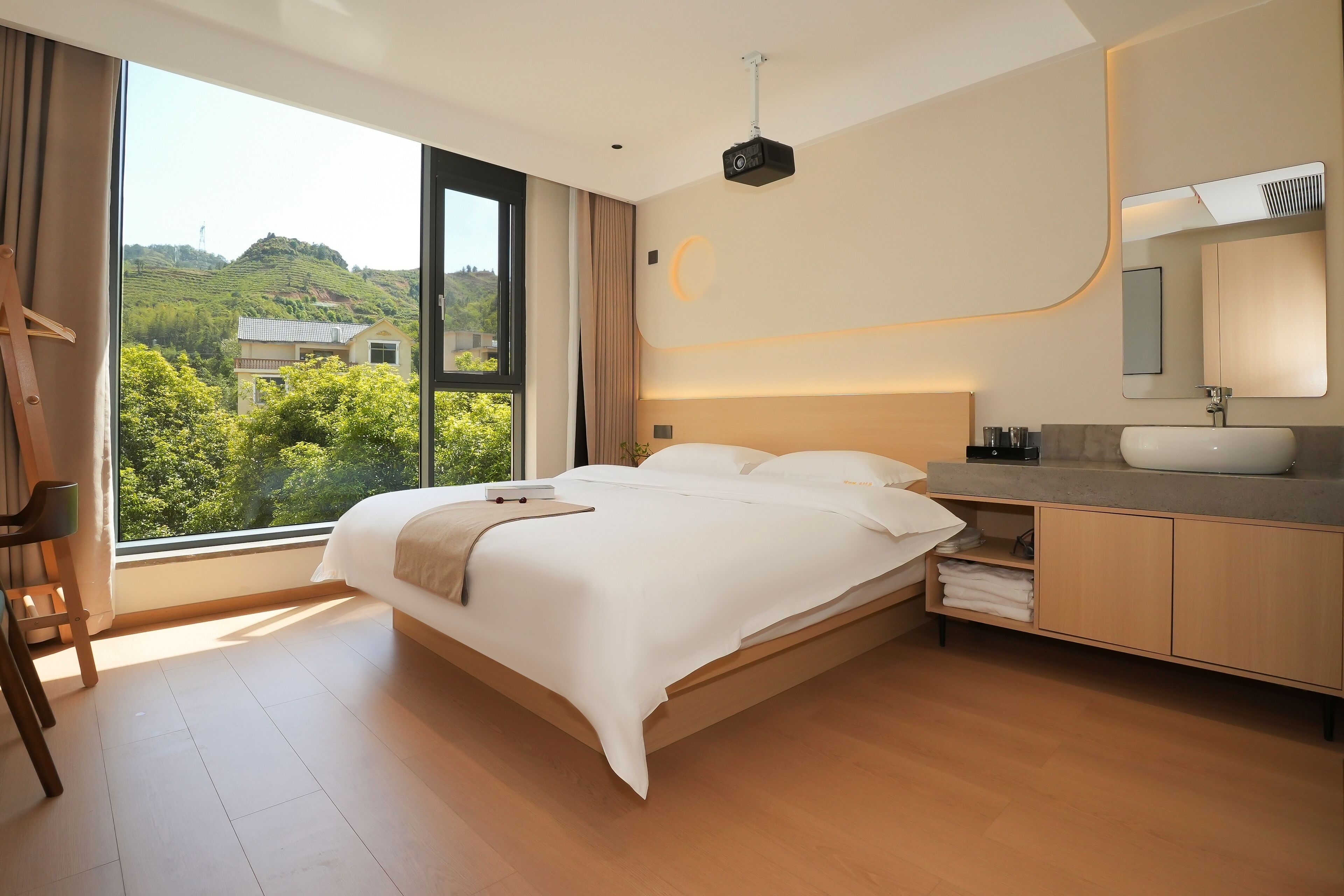 Ein modernes Hotelzimmer mit einem großen Bett, einem Nachttisch, einem Badezimmer mit Waschbecken und Spiegel sowie Blick auf Grünflächen und Hügel durch das Fenster.
