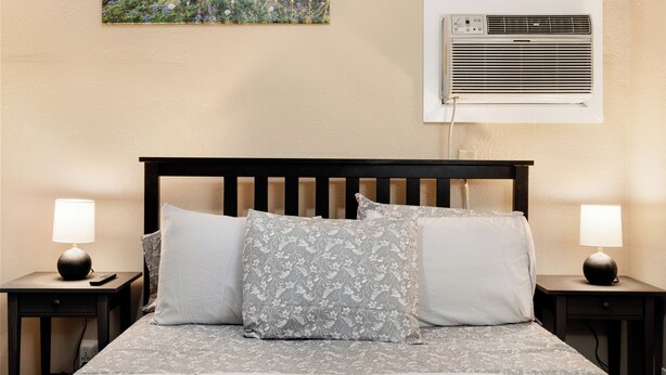 A neatly made bed with a floral-patterned pillow, a bedside lamp, and an air conditioning unit mounted on the wall. - Premium Cozy 1-bedroom apartment in fantastic Sacramento w/ AC and gym equipment (Sacramento)