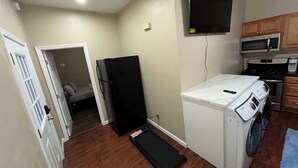 A compact kitchen with a microwave, stove, and washing machine. A black refrigerator is visible, and a TV is mounted on the wall. - Premium Cozy 1-bedroom apartment in fantastic Sacramento w/ AC and gym equipment (Sacramento)