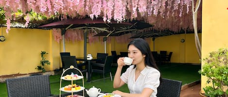 Una persona sentada a una mesa con un juego de té, rodeada de sillas y un pequeño jardín con césped artificial y plantas.