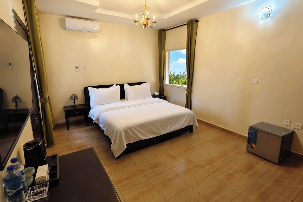 Habitación de hotel con cama, escritorio con lámpara, una mesita, un cesto de basura, una botella de agua, una caja y un pequeño calentador.