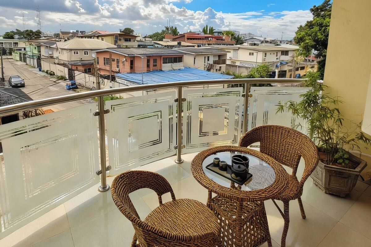 Un balcón con mobiliario de mimbre, una mesa de vidrio y vistas a una zona residencial con casas y una calle.