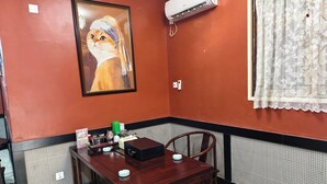 A dining area with a wooden table, chairs, a framed painting, an air conditioning unit, and a window with a lace curtain. - JUN YUE Ikeja Lagos hotel (Lagos)
