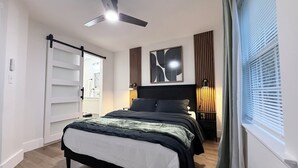 A modern bedroom with a bed, a ceiling fan, a bedside table with a lamp, a window with blinds, and a door leading to a bathroom.