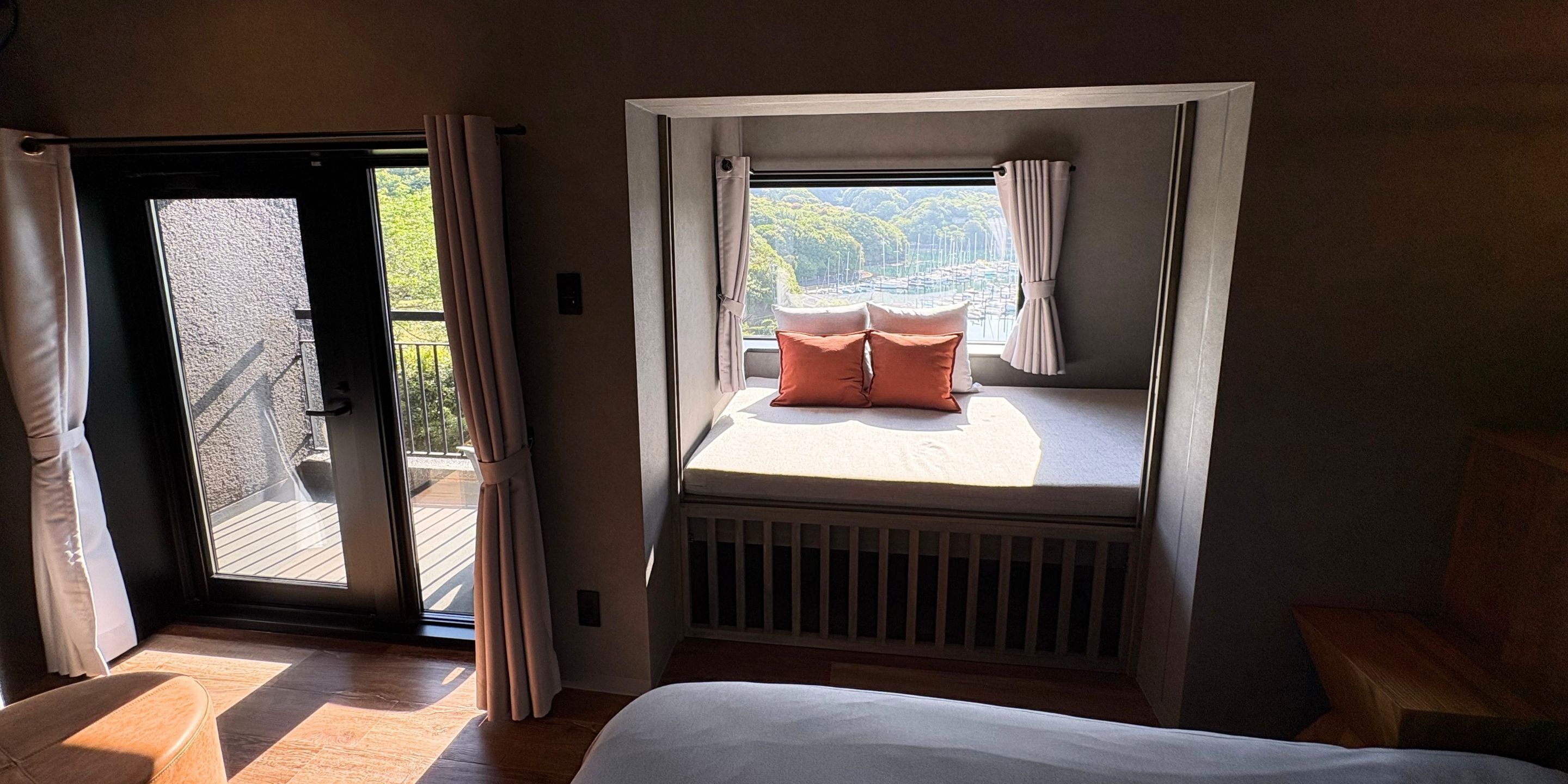 Ein Schlafzimmer mit Bett, einem Fenster mit Vorhängen, Blick ins Freie und einer Schiebetür, die auf einen Balkon führt.