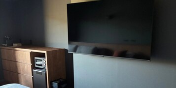 Habitación de hotel con televisor de pantalla plana montado en la pared, mueble de madera con microondas y refrigerador pequeño, y un cesto de basura.