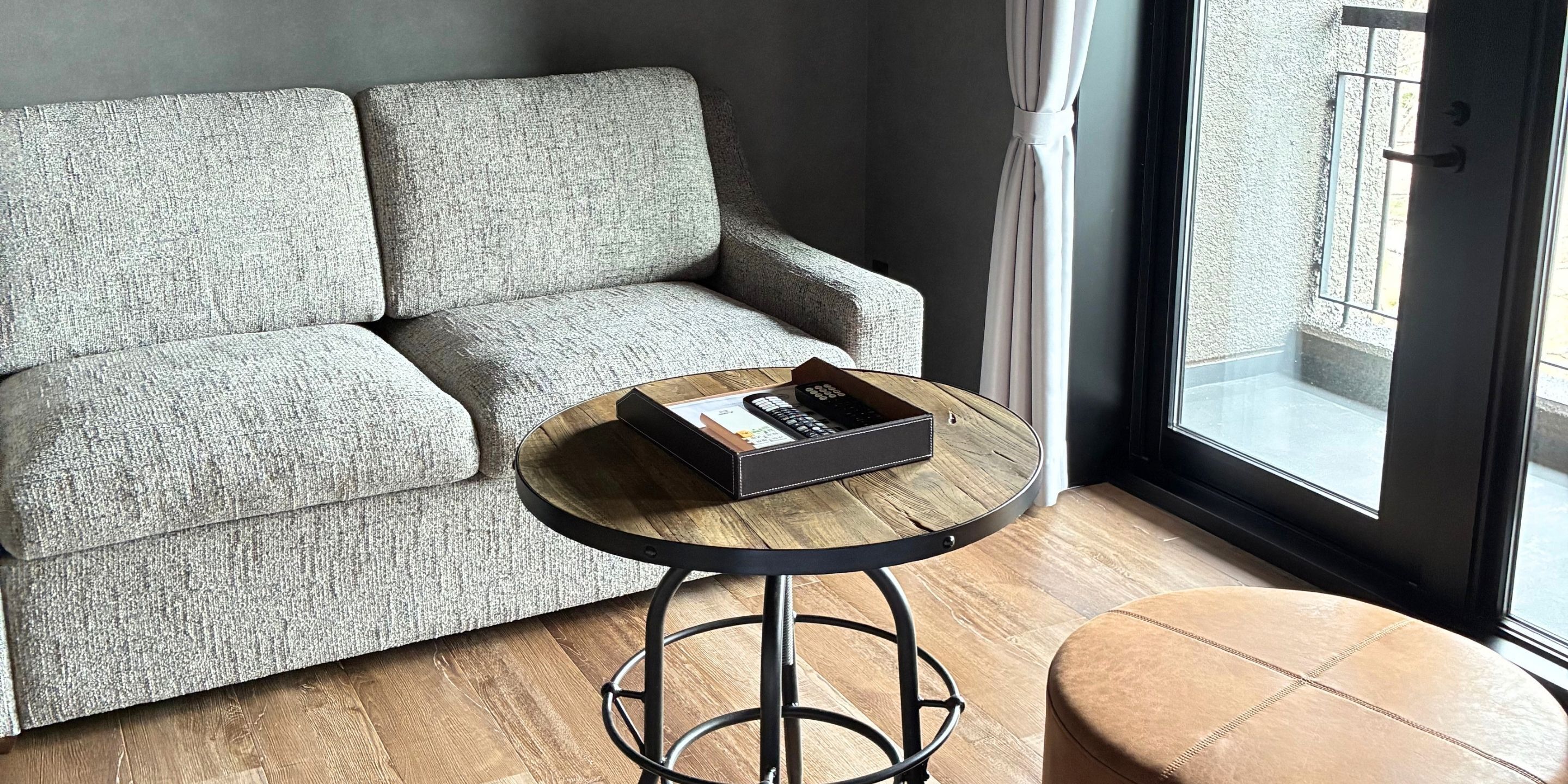 Un sofá gris, una mesa redonda de madera con base de metal negro y un otoman marrón en una habitación con un ventanal grande.