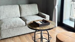 Un sofá gris, una mesa redonda de madera con base de metal negro y un otoman marrón en una habitación con un ventanal grande.