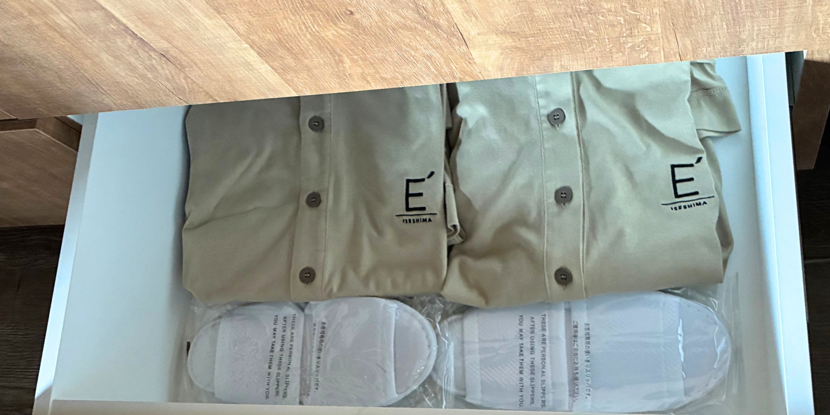 Un tiroir contenant quatre sacs beiges ornés d’un logo et d’un texte, ainsi que deux paires de chaussons blancs avec des instructions imprimées.