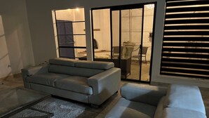 Ein modernes Wohnzimmer mit einem grauen Sofa, einem Glastisch und einer Glasschiebetür, die zu einer Außenpatio führt.