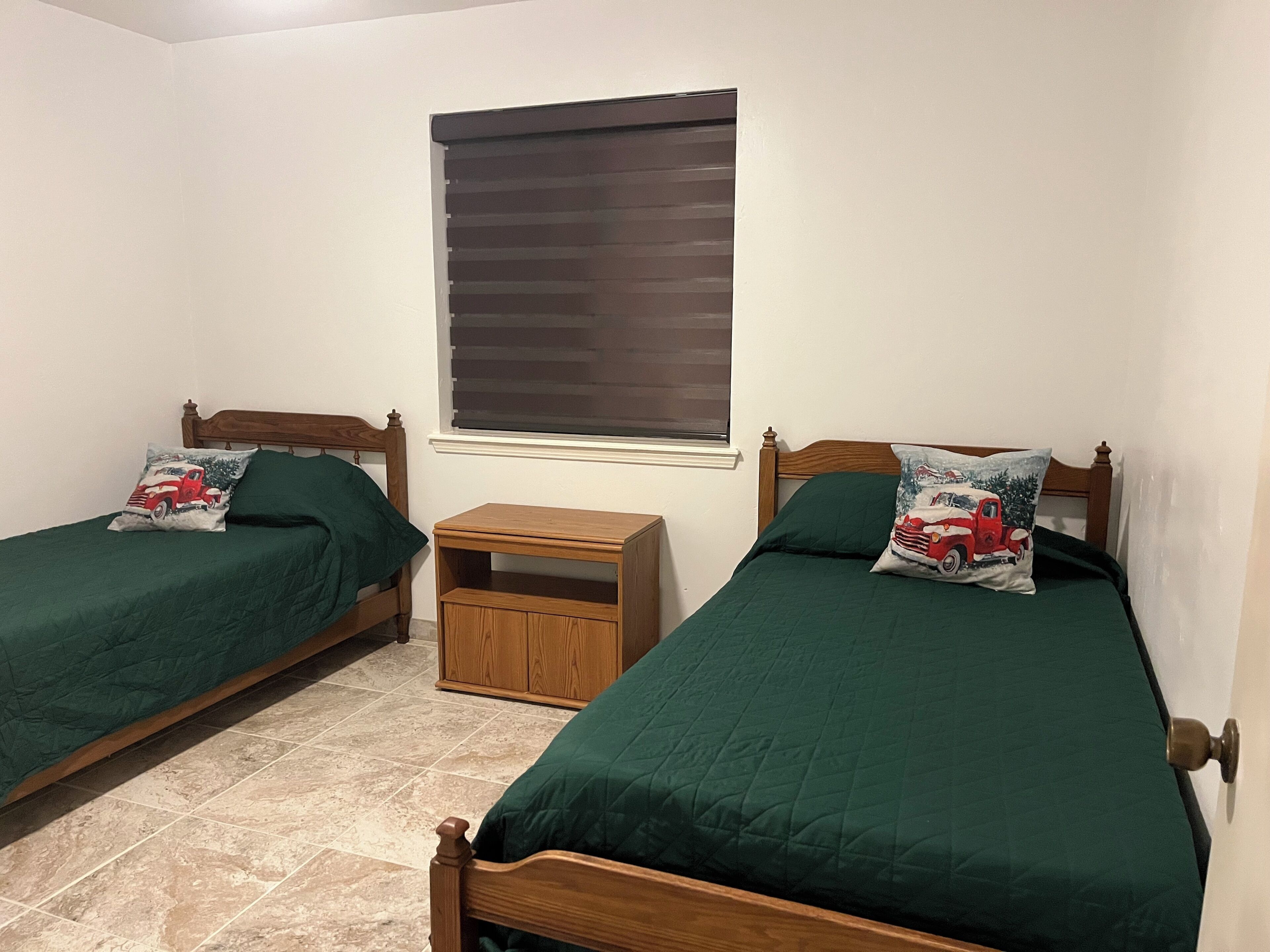 Un dormitorio con dos camas individuales, cada una con colcha y almohadas verdes a juego, una mesita de noche de madera y una ventana con persianas.