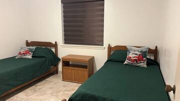 Un dormitorio con dos camas individuales, cada una con colcha y almohadas verdes a juego, una mesita de noche de madera y una ventana con persianas.