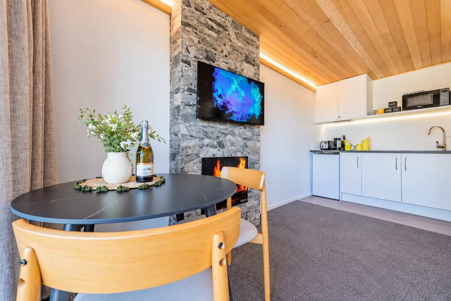 A modern living space with a round table, chairs, a fireplace, a television mounted on a stone wall, and a kitchen area with cabinets and appliances.