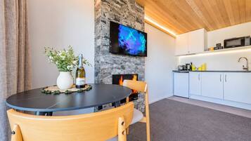 A modern living space with a round table, chairs, a fireplace, a television mounted on a stone wall, and a kitchen area with cabinets and appliances.