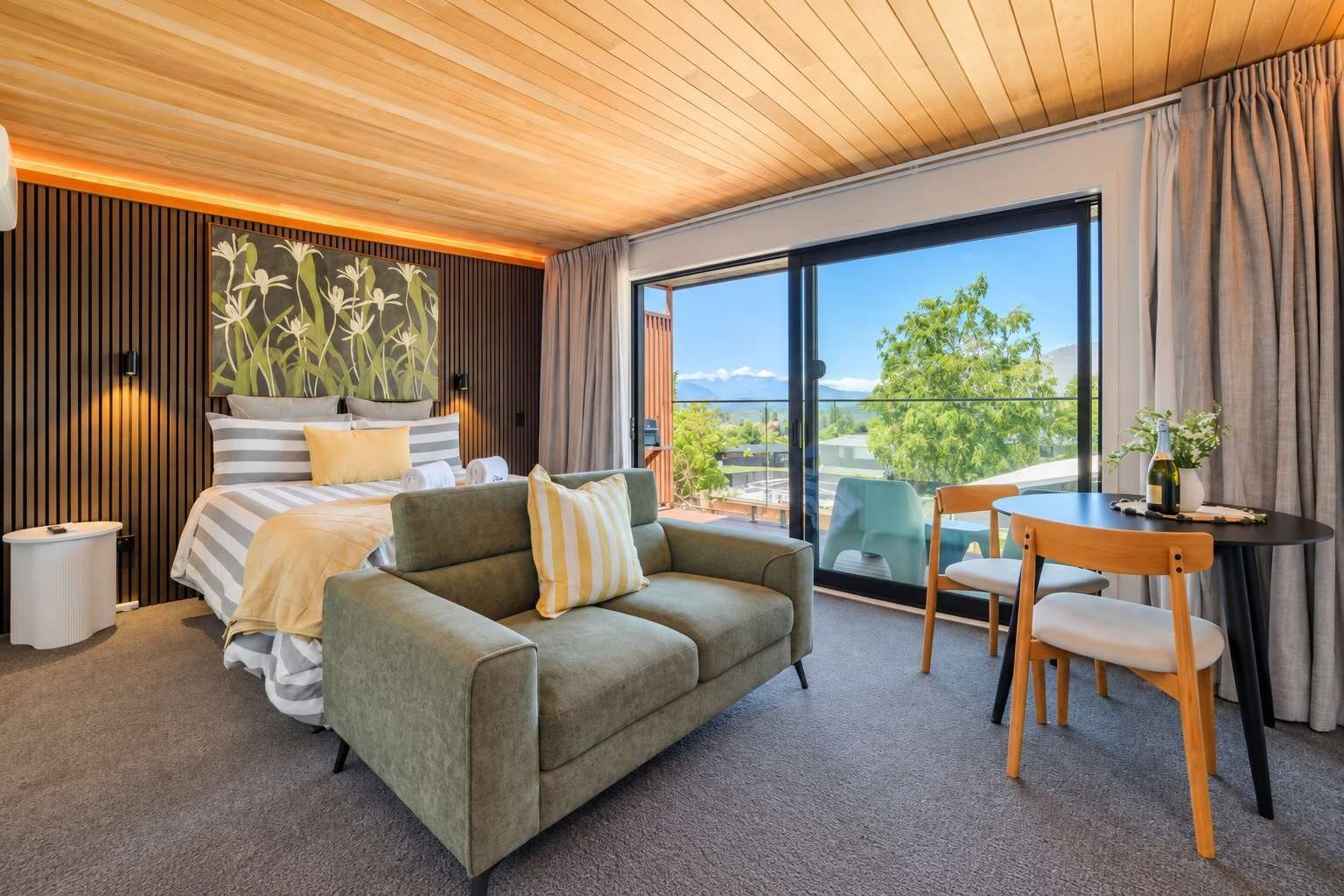 A modern hotel room with a bed, a sofa, a dining table, and a balcony with a view.