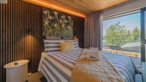 Ein modernes Hotelzimmer mit einem großen Bett, gestreifter Bettwäsche, einer Topfpflanze und Blick auf Grünflächen sowie eine Bahnstrecke.