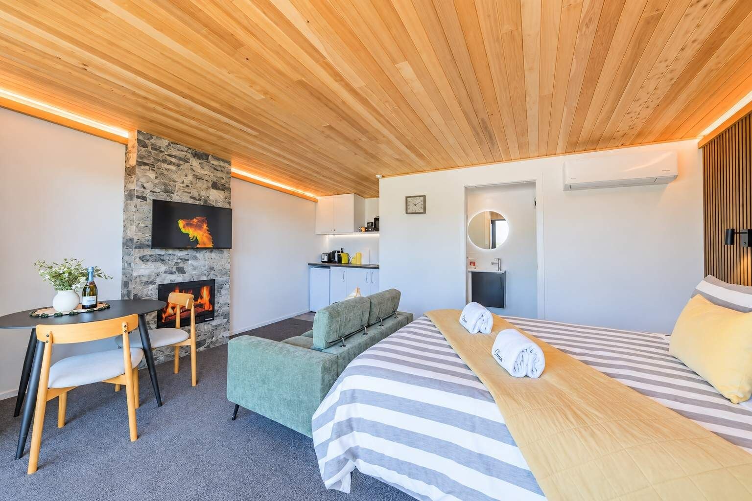 Una habitación moderna con chimenea, una cama con ropa de cama a rayas, un sofá, una mesa de comedor con sillas y una zona de cocina.