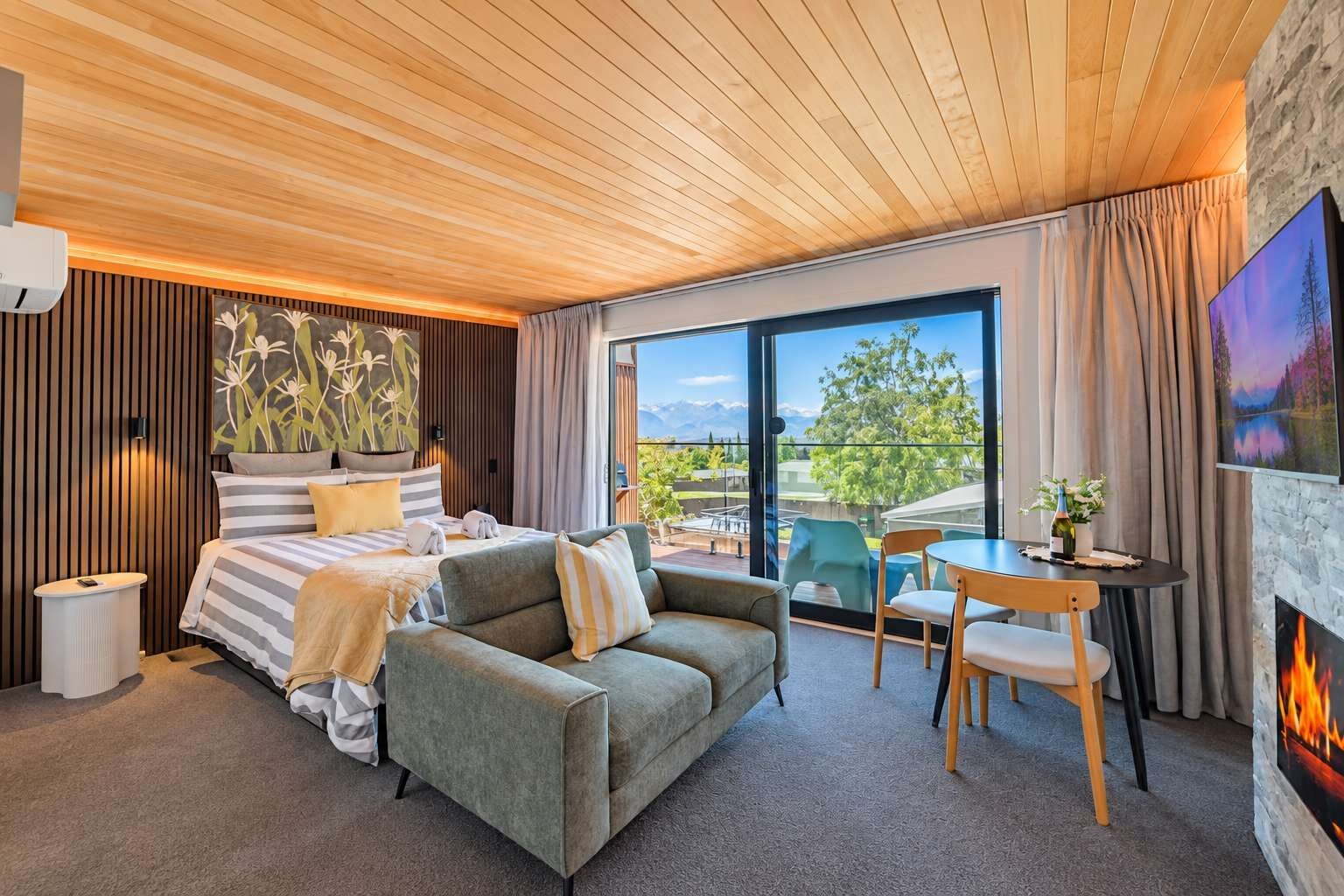 A modern hotel room with a bed, a sofa, a dining table, and a fireplace.