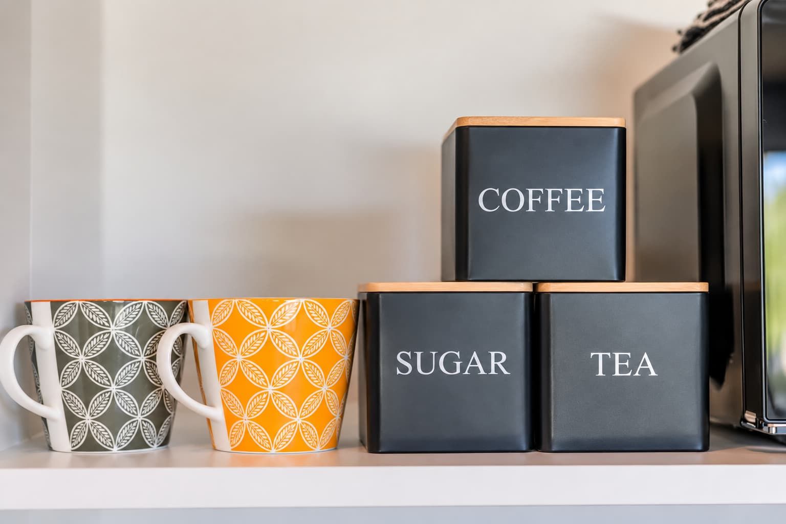 Une étagère avec trois récipients étiquetés « café », « sucre » et « thé », ainsi que deux tasses décorées.