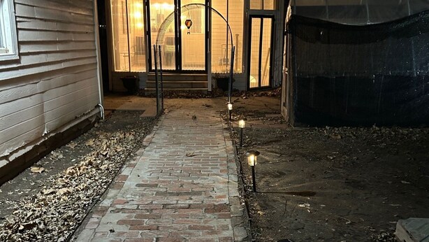Ein Ziegelweg führt zu einem Hauseingang mit überdachter Veranda und einem kleinen Gartenbereich mit dekorativer Beleuchtung.