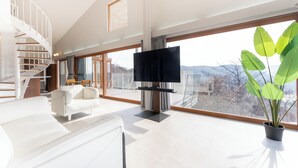 Ein modernes Wohnzimmer mit einem weißen Sofa, einem Flachbildfernseher, einer Treppe und großen Fenstern mit Blick ins Freie.