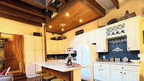 A kitchen with white cabinets, a marble countertop, and a wooden ceiling. There are stools at the island and a refrigerator on the right. - Luxurious Rooftop French Quarter Penthouse (New Orleans)